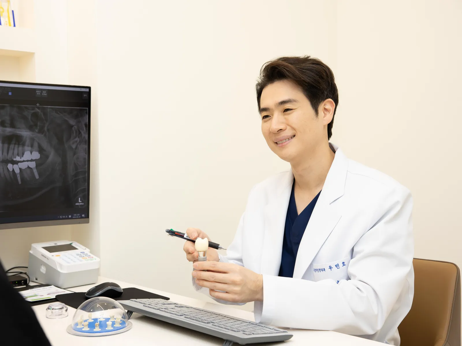 엑스레이 화면 옆에서 임플란트 모형을 들고 설명하는 우민호 대표원장